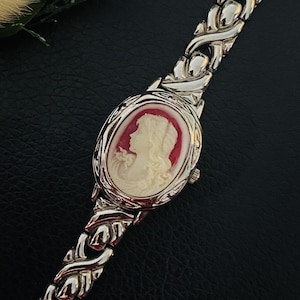 Puede incluir: Un reloj de pulsera plateado con una esfera ovalada que presenta un perfil de camafeo color crema de una persona sobre un fondo rojo oscuro. La correa del reloj está compuesta por eslabones metálicos plateados entrelazados con acabado pulido.