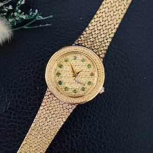 May include: A gold-tone wristwatch with a textured, woven-style metal band. The round watch face features a shimmering gold-colored dial with small green circular hour markers and gold hands. The brand name Klaeuse is printed on the dial.