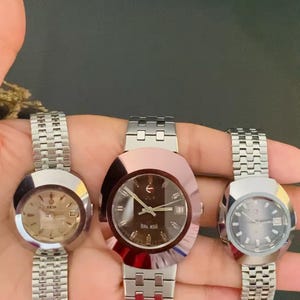 May include: Three Rado wristwatches with metallic oval-shaped cases and silver-toned link bracelets. The left watch has a light dial, the center watch features a dark brown dial with the text BALBOA, and the right watch has a dark grey dial.