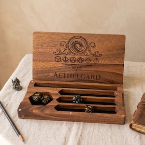 May include: A rectangular wooden dice storage box with a hinged lid featuring an engraved dragon emblem, geometric shapes, and the text AETHELGARD. The interior contains a hexagonal compartment and three horizontal slots holding dark dice.