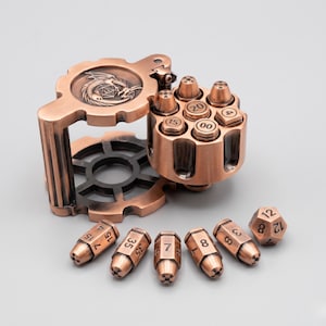 May include: A copper-coloured metal revolver cylinder dice holder with a hinged lid featuring a dragon emblem. The cylinder contains six bullet-shaped dice with numbers, and five additional bullet-shaped dice and one dodecahedron die are arranged in front.