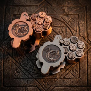 May include: Two metal dice holders shaped like revolver cylinders, one in copper and one in silver tone. Each features a hinged lid embossed with a dragon encircling a twenty-sided die. The cylinders have six circular slots containing numbered dice.