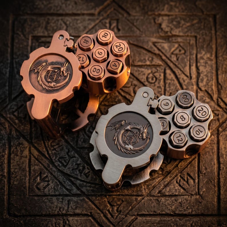 May include: Two metal dice holders shaped like revolver cylinders, one in copper and one in silver tone. Each features a hinged lid embossed with a dragon encircling a twenty-sided die. The cylinders have six circular slots containing numbered dice.