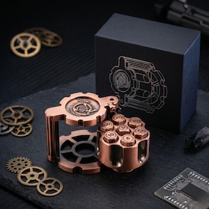 May include: A copper-coloured metal dice roller shaped like a revolver cylinder with numbered faces, featuring a dragon emblem on the top gear-shaped lid. It sits beside a black box with a matching gear and dragon line art design, surrounded by loose metal gears.