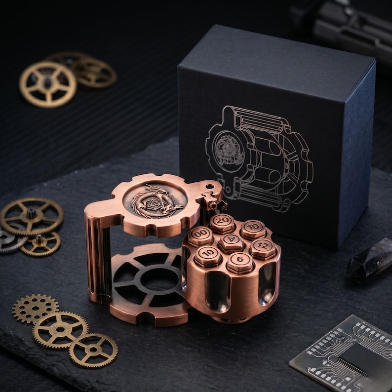 May include: A copper-coloured metal dice roller shaped like a revolver cylinder with numbered faces, featuring a dragon emblem on the top gear-shaped lid. It sits beside a black box with a matching gear and dragon line art design, surrounded by loose metal gears.