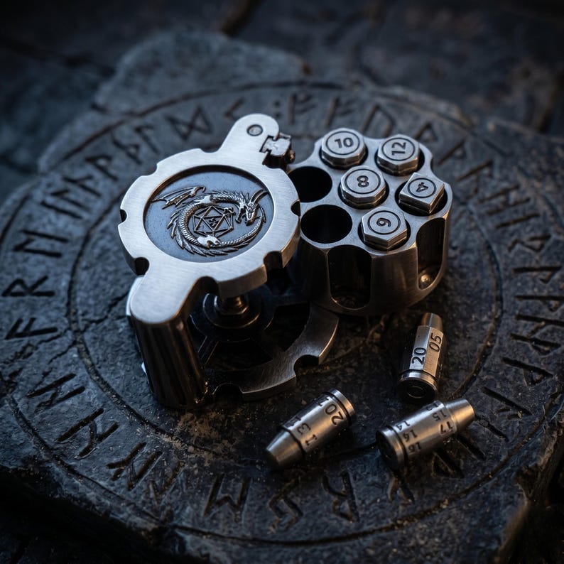 May include: A metallic silver revolver cylinder dice holder featuring a dragon and icosahedron emblem on the side. The cylinder contains several numbered dice inserts, and three bullet-shaped dice are positioned nearby on a dark stone surface.