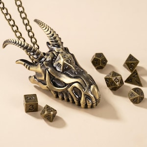 May include: A bronze-toned metal dragon skull pendant on a matching chain, featuring a twenty-sided die integrated into the forehead. The set includes six additional polyhedral dice in various shapes, all finished in a matching antique bronze colour.