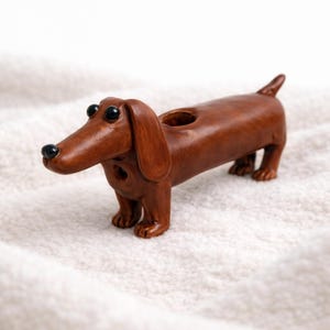 May include: A brown dachshund ornament with a hole in its back. The dog has black eyes and a black nose. The item is made of a smooth material and is standing on a white, textured surface.