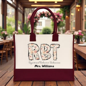 May include: A cream-colored canvas tote bag with a dark maroon base and matching handles. The front features the large letters RBT filled with a floral pattern, followed by the text Registered Behavior Technician and the name Mrs. Williams.