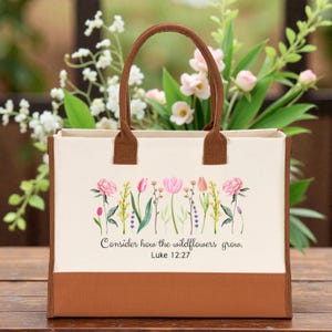 May include: A cream-colored canvas tote bag with brown trim and handles. The front features a watercolor illustration of pink and yellow wildflowers with green stems. Below the flowers, the text reads: Consider how the wildflowers grow. Luke 12:27.