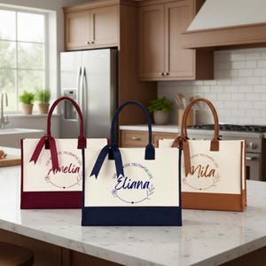 May include: Three rectangular canvas tote bags with top handles and matching ribbons. Each bag features a name in cursive script, a floral wreath graphic, and the text THE TRUTH THE LIFE. The bags are colored in burgundy, navy blue, and brown.
