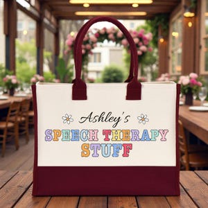 May include: A cream-colored canvas tote bag with dark maroon handles and a matching maroon base. The front features the text "Ashley's SPEECH THERAPY STUFF" in colorful, blocky letters, flanked by two small white daisy illustrations on either side.