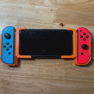 May include: A gaming device with a black screen, flanked by a blue controller on the left and a red controller on the right. The device is held in place by an orange frame, set against a wooden surface.