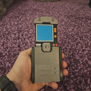 Puede incluir: Un dispositivo portátil gris con una pantalla azul y botones naranjas. El dispositivo tiene forma rectangular con una tapa con bisagras y un borde negro. El dispositivo se sostiene en una mano, con un reloj negro visible en la muñeca.