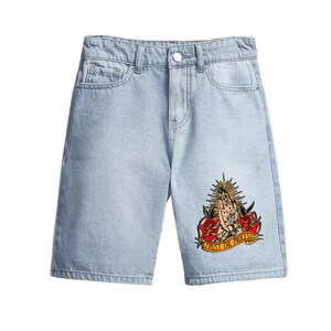 May include: Pale blue denim shorts with a classic five-pocket design. The shorts have a colourful graphic on the right leg featuring praying hands, roses, and the text "TRUST THE PROCESS".