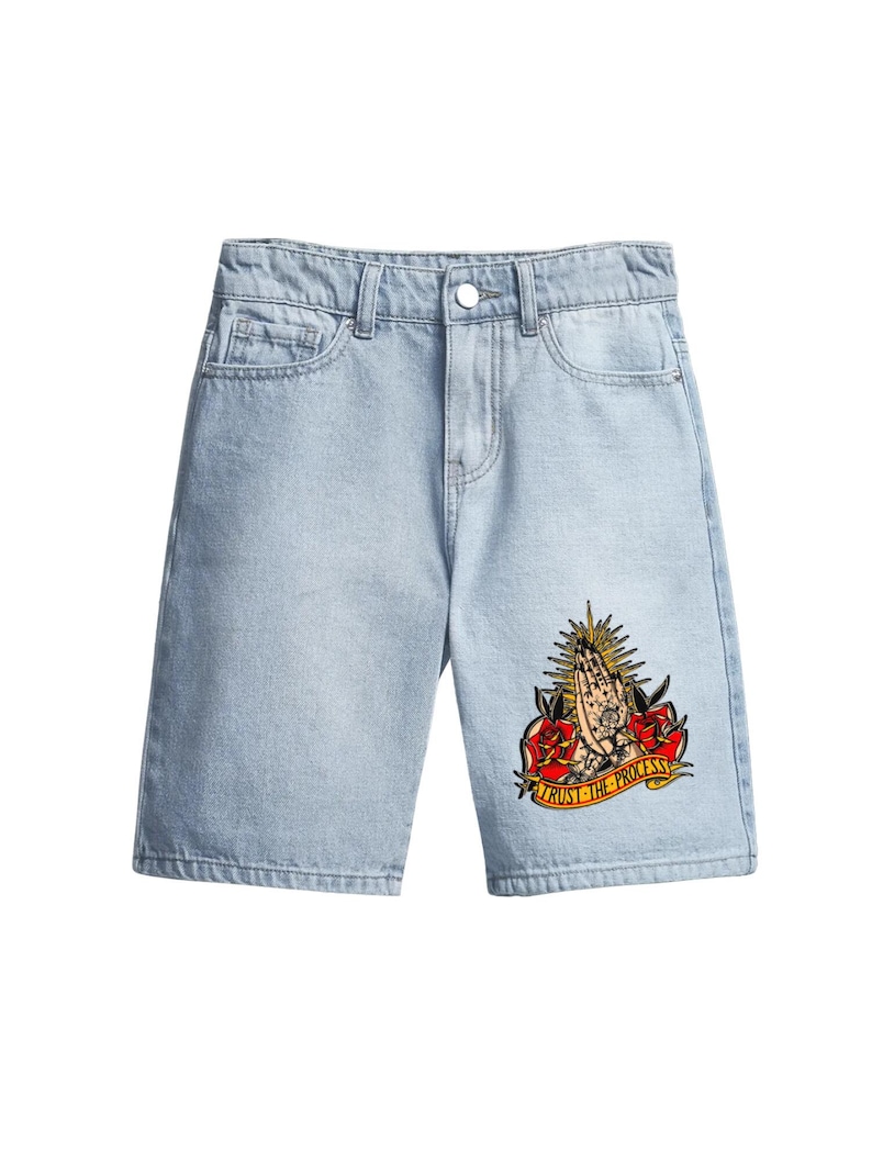 May include: Pale blue denim shorts with a classic five-pocket design. The shorts have a colourful graphic on the right leg featuring praying hands, roses, and the text "TRUST THE PROCESS".