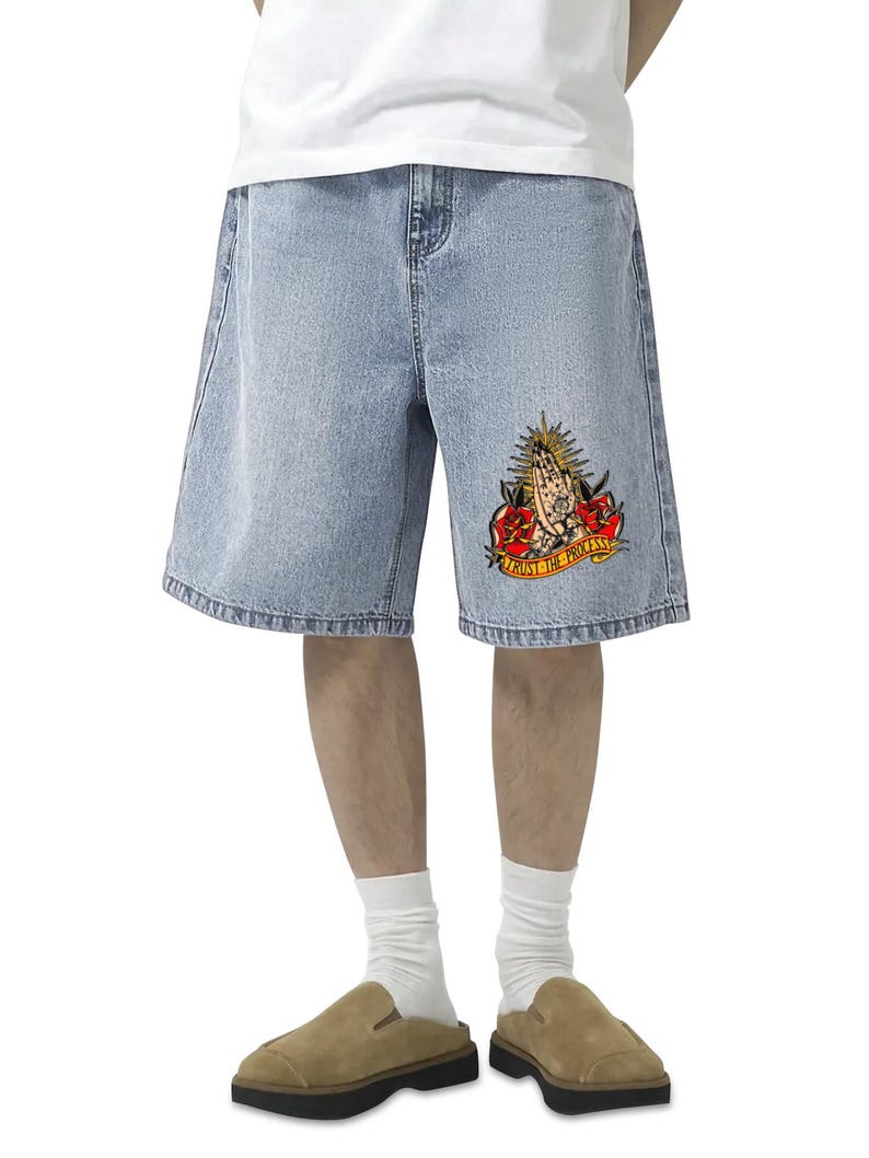 May include: Light blue denim shorts with a relaxed fit. The shorts feature a colorful graphic on the right leg with praying hands, roses, and the text "TRUST THE PROCESS". The shorts are worn with a white t-shirt, white socks, and brown suede slip-on shoes.