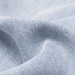 May include: Close-up of light blue denim fabric with a diagonal weave pattern. The fabric is slightly creased, creating shadows and highlights. The texture suggests a durable and comfortable material, suitable for clothing.