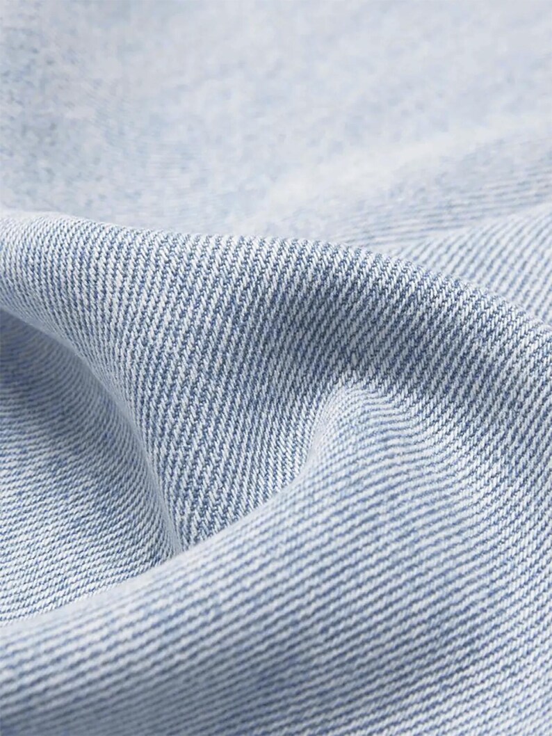 May include: Close-up of light blue denim fabric with a diagonal weave pattern. The fabric is slightly creased, creating shadows and highlights. The texture suggests a durable and comfortable material, suitable for clothing.