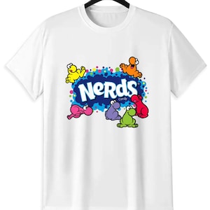 May include: White t-shirt featuring a colorful graphic of the word "Nerds" in blue, surrounded by cartoon characters in yellow, orange, pink, purple, green, and red. The shirt is hanging on a black hanger.