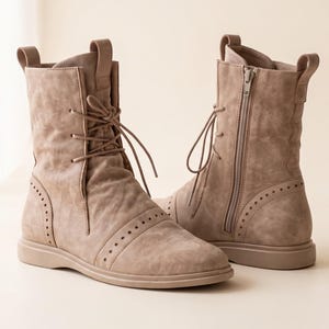 May include: A pair of taupe mid-calf boots featuring a lace-up front closure, a side zipper, and decorative perforated detailing along the toe cap and heel. The boots have a flat sole, a pull tab at the back, and a smooth, matte finish throughout.