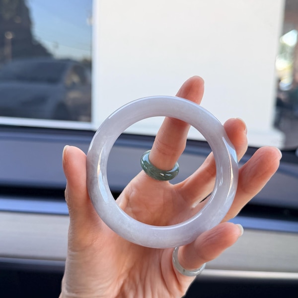 May include: A smooth, translucent lavender jade bangle bracelet with a polished finish, paired with a smaller, dark green jade ring worn on the finger, showcasing natural color variations and a rounded, circular profile.