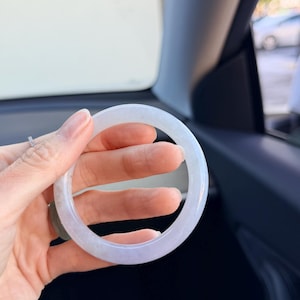 May include: A smooth, circular bangle bracelet crafted from translucent, pale lavender-toned stone. The material exhibits a subtle, cloudy texture with soft white and light purple color variations throughout the polished, rounded surface.