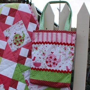 May include: A pink and green patchwork quilt with a red and white polka dot pattern. A small tote bag with a pink and green floral print and a red scalloped trim is hanging on a white picket fence.