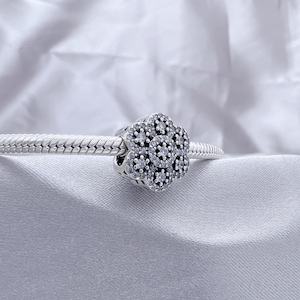 Könnte beinhalten: Ein silberner Charm mit einem Blumendesign, besetzt mit kleinen, klaren Edelsteinen. Der Charm befindet sich auf einem silbernen Armband, vor einem weißen, strukturierten Hintergrund. Der Blumen-Charm ist ein Schmuckzubehör.