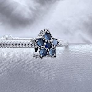 Puede incluir: Un charm de plata con diseño de estrella, con un grupo de piedras preciosas azules. La estrella está engarzada en una pulsera de plata con un patrón trenzado texturizado. Las piedras preciosas varían en tonos azules.