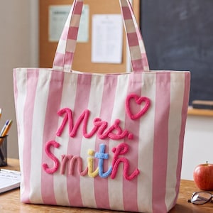 May include: A pink and white striped canvas tote bag featuring the embroidered text Mrs. Smith in raised pink, yellow, and blue yarn lettering, accompanied by a small pink yarn heart above the text and two matching striped shoulder straps.