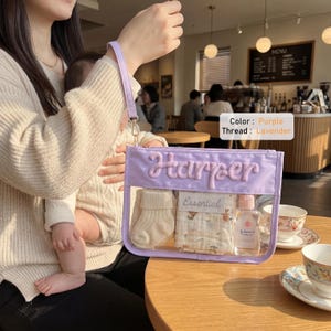 May include: A rectangular lavender pouch with a clear front panel and a wrist strap. The name Harper is embroidered in lavender thread on the front. Inside the pouch are a pair of cream-colored knit socks, a patterned cloth, and a small bottle.
