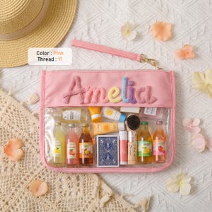 May include: A pink rectangular pouch with a clear front panel and a wrist strap. The name Amelia is embroidered in multi-colored yarn on the top section. The pouch contains various miniature bottles, a deck of cards, and small tubes.