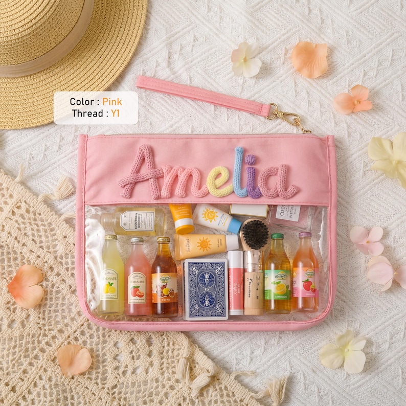 May include: A pink rectangular pouch with a clear front panel and a wrist strap. The name Amelia is embroidered in multi-colored yarn on the top section. The pouch contains various miniature bottles, a deck of cards, and small tubes.