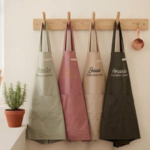 Pode incluir: Quatro aventais personalizados pendurados em um cabide de madeira. Os aventais são em tons de verde, rosa, bege e verde escuro. Cada avental tem um nome e uma frase impressa. Um pequeno vaso com uma planta e uma xícara de medição de cobre também são visíveis.