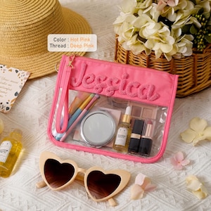 May include: A hot pink rectangular clear pouch with a wrist strap, featuring the name Jessica embroidered in deep pink thread. The pouch contains makeup brushes, a round compact mirror, a small bottle, and two lipsticks. Heart-shaped sunglasses sit below.