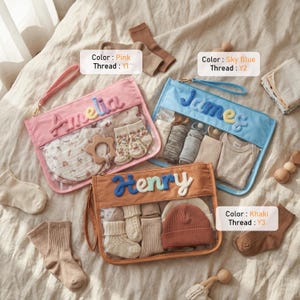 May include: Three rectangular zippered pouches in pink, sky blue, and khaki, each featuring a clear front panel and a personalized name in textured yarn: Amelia, James, and Henry. The pouches contain baby items like socks, hats, and bibs.