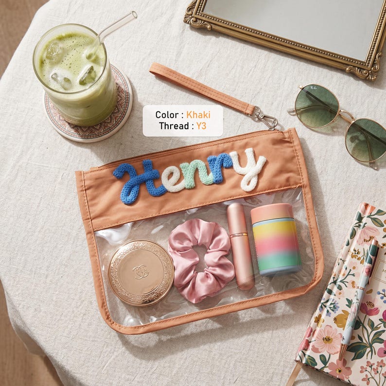 May include: A khaki pouch with a clear plastic front panel and a wrist strap. The name Henry is embroidered in blue, light green, and white yarn across the top. Inside are a gold compact, a pink satin scrunchie, a lipstick, and a colorful cup.