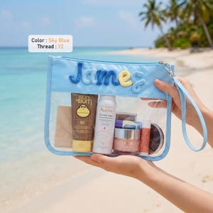 May include: A sky blue transparent pouch with a wrist strap, featuring the name James embroidered in blue and yellow yarn. The pouch contains a Sun Bum sunscreen tube, an Avene thermal water spray, a Clinique jar, and other skincare items.