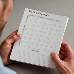 May include: A white Week Meal Prep notepad with a grid layout for daily meal planning. The notepad includes sections for breakfast, lunch, dinner, and snacks, with a grocery list at the bottom. The notepad is held by a person.