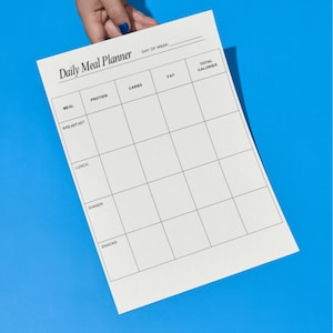 Puede incluir: Una hoja blanca de Daily Meal Planner con secciones para comida, proteínas, carbohidratos, grasas y calorías totales. El planificador se sostiene contra un fondo azul brillante. El texto "Daily Meal Planner" es visible.