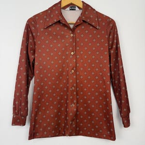 May include: A long-sleeved button-down shirt in a deep rust-brown color featuring a repeating geometric pattern of small white diamonds with dark centers. The shirt has a pointed collar, a front button placket, and buttoned cuffs on the sleeves.