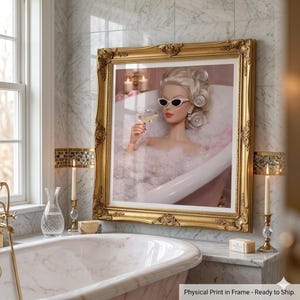 May include: A framed print of a doll in a bathtub, wearing sunglasses and holding a glass of wine. The gold frame is ornate, and the print is displayed in a marble bathroom with candles and a vintage bathtub.