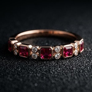 May include: A rose gold ring featuring a series of square-cut red gemstones alternating with small, round, clear gemstones. The ring is set against a dark, textured background, highlighting the sparkle of the gems.
