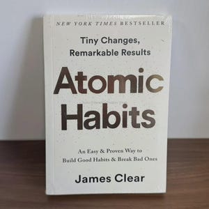 May include: A white book titled "Atomic Habits" by James Clear. The cover features the title in large, bold letters, with the subtitle "Tiny Changes, Remarkable Results." The book is a New York Times Bestseller, and it is wrapped in clear plastic.