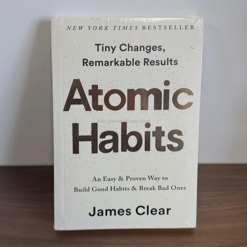 May include: A white book titled "Atomic Habits" by James Clear. The cover features the title in large, bold letters, with the subtitle "Tiny Changes, Remarkable Results." The book is a New York Times Bestseller, and it is wrapped in clear plastic.