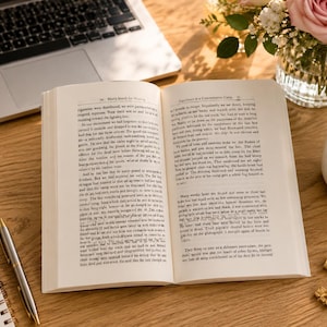 May include: An open book with text, a laptop, a pen, a notepad, a cup of coffee, a vase of flowers, and gold binder clips are arranged on a wooden surface. The book is open to a page of text, and the scene is lit by natural light.