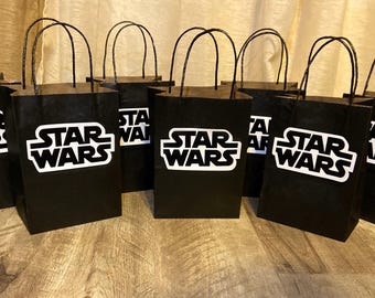 May include: A collection of seven black paper gift bags, each featuring the white Star Wars logo printed on the front. The bags have black twisted paper handles and are arranged in a group, displaying the iconic branding clearly on each unit.