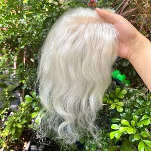 May include: A hair topper featuring long, wavy, platinum blonde strands with a visible lace base at the crown. The hair has a soft, natural texture and a light, bright colour tone, designed to provide coverage and volume for the top of the head.