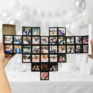 Op de afbeelding: Een hartvormig fotoalbum met meerdere kleine ingelijste foto's. Het album is open en toont talrijke foto's. De achtergrond omvat witte ballonnen, een taart en geschenkdozen, wat duidt op een viering of speciale gelegenheid.